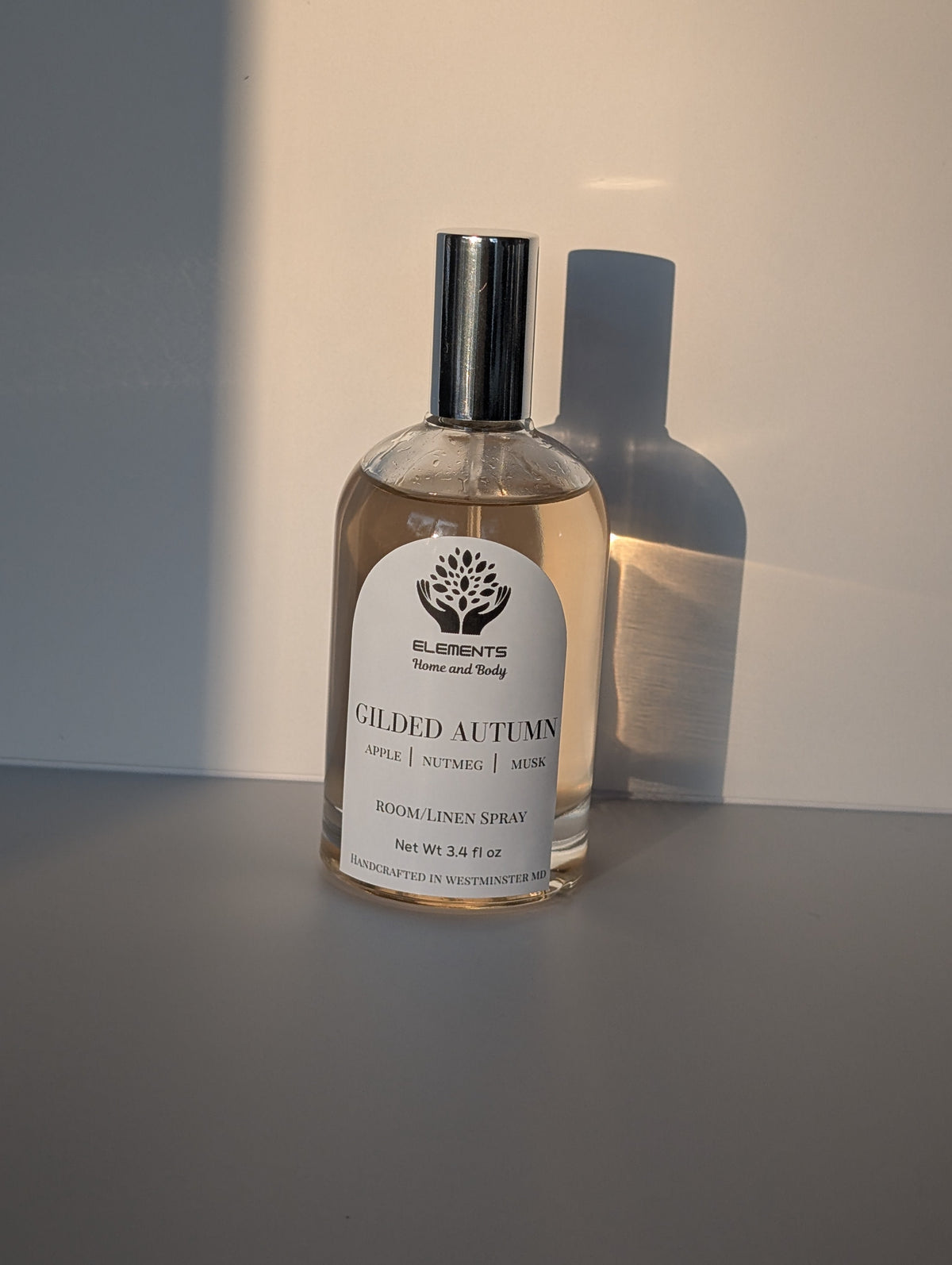 A bottle of Elements Trees and Body 'Gilded Autumn' linen spray, featuring a clear glass bottle with a silver cap and a white label, set against a warm, sunlit beige background with a soft shadow.